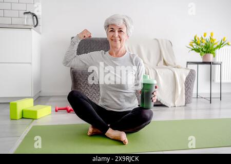 Fröhliche Ältere Frau Schaut In Die Kamera Und Zeigt Ihren Bizeps, Der Einen Aktiven Lebensstil Und Heimtrainings Verkörpert Stockfoto