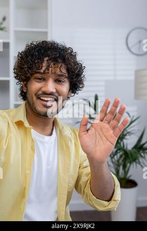 Ein lächelnder junger Mann winkt bei einem Videochat hallo, in einer modernen Inneneinrichtung. Warmes und freundliches Kommunikationskonzept. Stockfoto