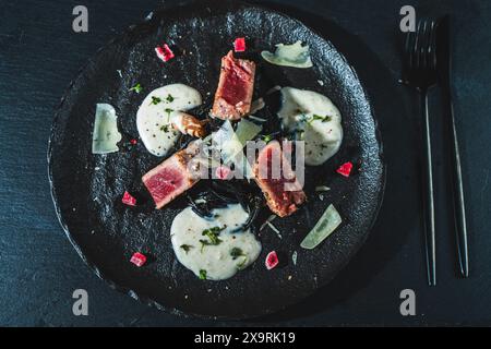 Gekochtes saftiges Thunfischsteak mit Gemüse und Tintenfisch-Tintenspaghetti auf einem schwarzen Steinteller. Essen im Restaurant. Fisch Und Meeresfrüchte. Country-STYLE. Flaches Layout. Hg Stockfoto