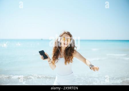 Lächelnde, moderne Frau am Meer mit Smartphone tanzen und Musik mit Kopfhörern hören. Stockfoto