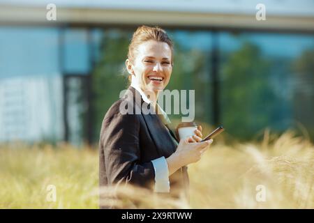 Porträt einer glücklichen modernen Arbeiterin in der Nähe des Bürogebäudes in schwarzer Jacke mit Smartphone und Tasse Kaffee. Stockfoto