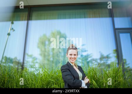 Lächelnde moderne Arbeiterin mittleren Alters im Geschäftsviertel in schwarzer Jacke mit überkreuzten Armen. Stockfoto