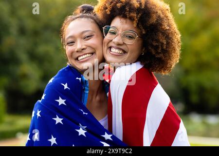 Porträt glücklicher, vielfältiger Freundinnen, bedeckt mit der Flagge der usa im Garten Stockfoto