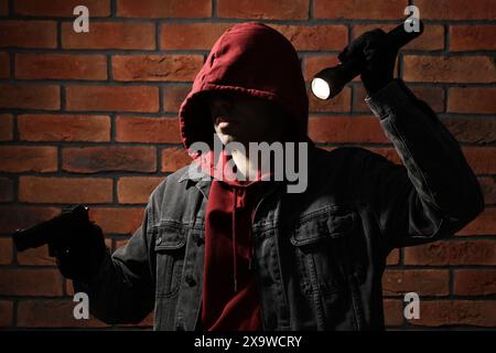 Dieb im Hoodie mit Pistole und Taschenlampe an der roten Ziegelwand Stockfoto