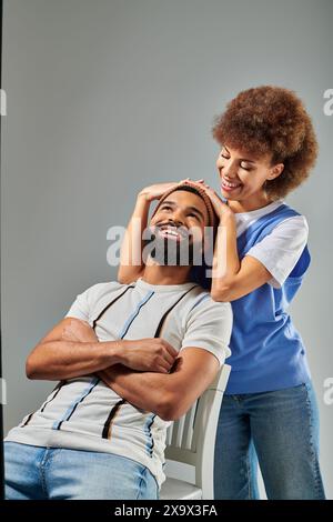 Ein afroamerikanischer Mann und eine afroamerikanische Frau in trendigen Outfits, die auf einer weißen Bank vor grauem Hintergrund sitzen. Stockfoto