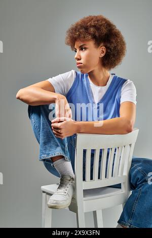 Ein junger Mann in stilvoller Kleidung sitzt anmutig auf einem weißen Stuhl vor grauem Hintergrund. Stockfoto