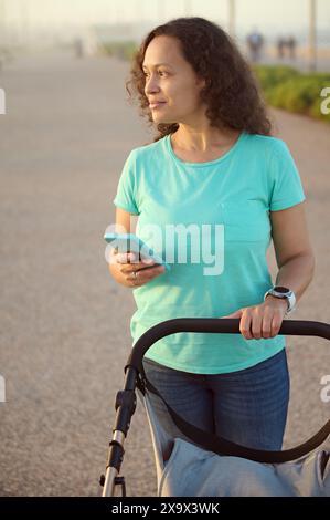 Vertikale Aufnahme einer lockigen, multiethnischen glücklichen jungen Frau 40er, liebevolle Mutter, die Babywagen schiebt, lächelnd, zur Seite schauend, auf dem Fußweg auf dem p Stockfoto