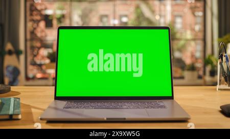 Laptop-Computerdisplay mit nachgeordnetem grünem Bildschirm auf einem Holzschreibtisch im Wohnzimmer. Chroma Key Monitor zu Hause oder im Creative Loft Office mit Notizblock, Maus und Stift Stockfoto