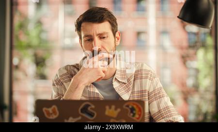 Porträt eines gutaussehenden Mannes, der an einem Laptop arbeitet, während er im stilvollen, gemütlichen Loft-Wohnzimmer sitzt. Freiberuflicher Mitarbeiter, Der Von Zu Hause Aus Arbeitet. Denken, Im Internet Surfen, Soziale Netzwerke Nutzen, Produktiv Sein Stockfoto