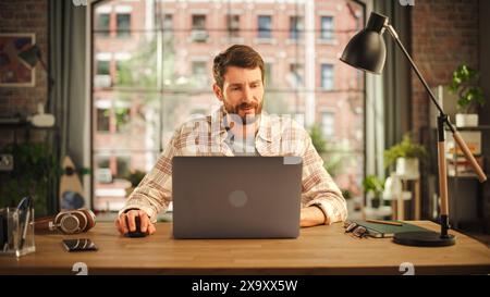 Kreativer Bärenmann, der an einem Laptop arbeitet, während er an einem Tisch im stilvollen gemütlichen Loft-Wohnzimmer sitzt. Freiberuflicher Mitarbeiter, Der Remote Arbeitet. Im Internet surfen, soziale Netzwerke nutzen, Projekte nachdenken. Stockfoto