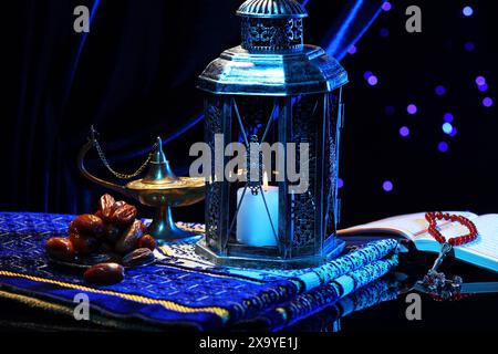 Arabische Laterne, Koran, misbaha, Aladdin Zauberlampe, Datteln und gefaltete Gebetsmatte auf Spiegeloberfläche vor verschwommenem Licht bei Nacht Stockfoto