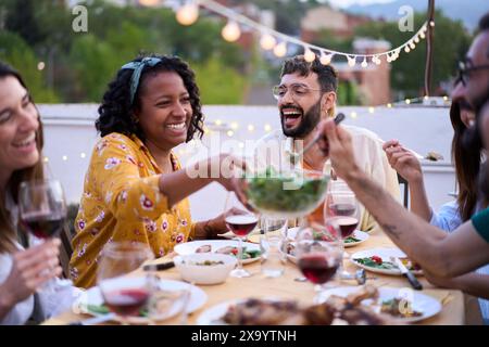 Lächelnde afroamerikanische Frau, die am Tisch sitzt und fröhlichen Freunden Salat bei einer Mahlzeit im Freien überreicht Stockfoto