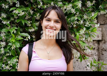 Niedliches Studentenmädchen in rosa T-Shirt und Rucksack, das die Kamera vor einer Wand lächelt, mit blühendem Jasmin und vom Wind verwobenen Haaren Stockfoto