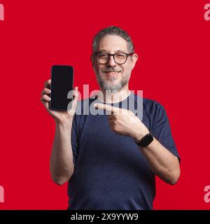 Reifer Mann mit Brille und Bart, lächelnd und auf einen Smartphone-Bildschirm zeigt - trägt ein blaues Hemd und steht vor einem leuchtend roten Hintergrund Stockfoto