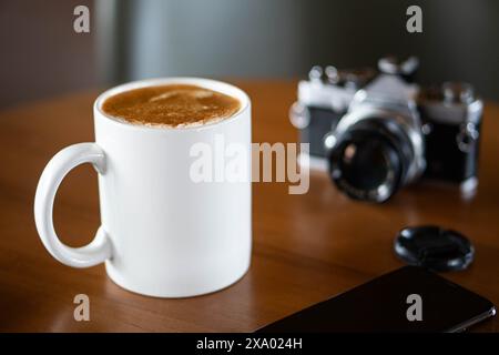 Eine alte Filmkamera auf einem Holztisch neben einer weißen Kaffeetasse. Die Kamera hat ein schwarzes und silbernes Gehäuse mit einer Objektivkappe in der Nähe. Stockfoto