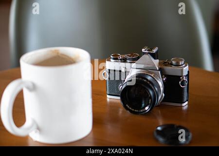 Eine alte Filmkamera auf einem Holztisch neben einer weißen Kaffeetasse. Die Kamera hat ein schwarzes und silbernes Gehäuse mit einer Objektivkappe in der Nähe. Stockfoto