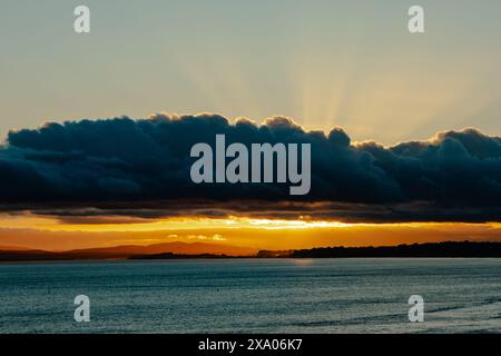 Die dunkelblauen Wolken mit orangefarbenem Sonnenuntergang erzeugen Strahlen aus den Wolken vom Strand mit Blick auf das Meer. Stockfoto
