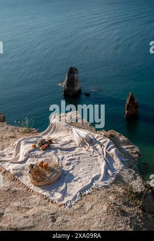 Eine weiße Decke ist an einem Strand in der Nähe des Wassers verteilt. Die Decke ist auf einem Felsen und es gibt ein paar Orangen und Äpfel darauf. Die Szene ist friedlich Stockfoto