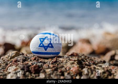 Israelische Flagge gemalt. Handgemalter Stein mit Magen David. Blaues Meer mit Wellen im Hintergrund Stockfoto