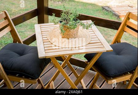 Blick aus nächster Nähe auf hölzerne Gartenmöbel auf einem kleinen Balkon. Rosmarin-Topf auf dem Tisch, natürliche Platzmatte. Stockfoto