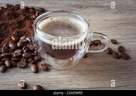 Glasbecher mit amerikanischem Kaffee, umgeben von Kaffeebohnen und gemahlenem Kaffee auf Holzbrett Stockfoto