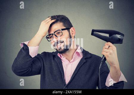 Junger, gutaussehender Mann in rosa Hemd und Brille, der Haare mit Haarfön pflegt und selbstbewusst aussieht. Stockfoto