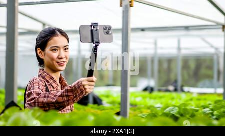 Eine lächelnde asiatische Frau, die in einem Gewächshaus arbeitet und mit ihrem Handy ein Selfie macht Stockfoto