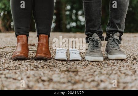 Zukünftige Mutter und Vater stehen neben weißen Baby-Sneakers während eines Mutterschaftsshootings im Park Stockfoto