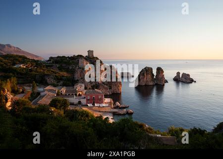 Ein wunderschöner Strand mit einem kleinen roten Gebäude in der Ferne bei Sonnenaufgang. Das Wasser ist ruhig und der Himmel ist wunderschön blau. Tonnara di Scopello, Provin Stockfoto