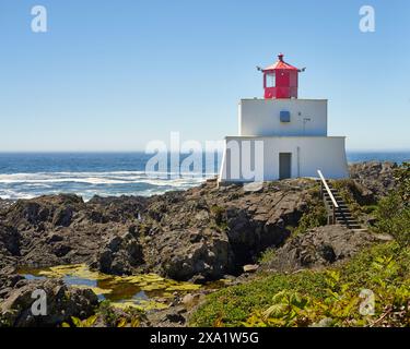 Amphitrite Point Lighthouse liegt hoch auf zerklüfteten braunen Felsen mit grüner Vegetation im Vordergrund und einem tiefblauen Ozean im Hintergrund. Stockfoto