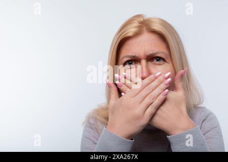 Eine Frau mittleren Alters mit blonden Haaren in einem grauen Rollkragen ist besorgt und bedeckt Gesicht und Mund mit ihren Händen Stockfoto