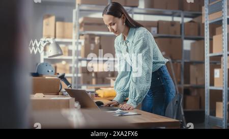 Die Eigentümerin eines Online-Shops arbeitet an einem Laptop, während sie an ihrem Schreibtisch im Lager steht. Weibliche Mitarbeiterin, die einen stilvollen gelben Pullover im Zimmer mit Regalen voller Pakete verpackt. Stockfoto