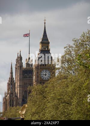 Ein hoher Uhrenturm mit britischer Flagge an einem bewölkten Himmel, umgeben von grünen Bäumen, London, England, Vereinigtes Königreich Stockfoto