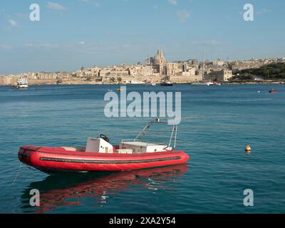 Ein rotes Schlauchboot schwimmt ruhig auf dem Meer vor der Stadt mit historischen Gebäuden im Hintergrund, Valetta, Malta Stockfoto