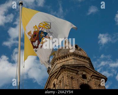 Gelbe und weiße Flagge mit Symbolen, die im Wind winken, vor einem historischen Gebäude und blauem Himmel, Valetta, Malta Stockfoto