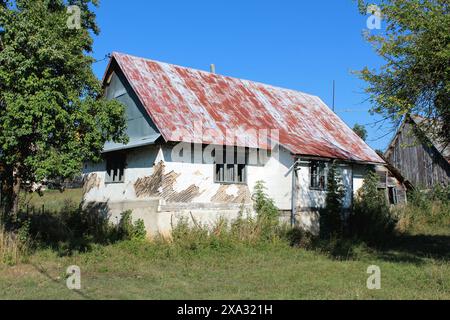Verlassenes kleines altes urbanes Familienhaus mit zerstörter weißer Fassade um baufällige Holzrahmenfenster mit Glasbruch, die mit Rot bedeckt sind Stockfoto
