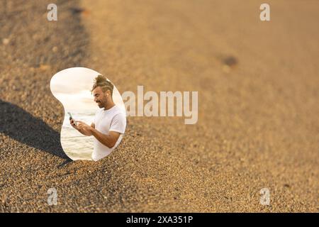 Mann mit Handy reflektiert in einem Spiegel am Strand mit sandigem Meer und Sonnenuntergang im Hintergrund Stockfoto