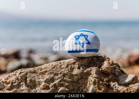 Israelische Flagge gemalt. Handgemalter Stein mit Magen David. Blaues Meer mit Wellen im Hintergrund Stockfoto