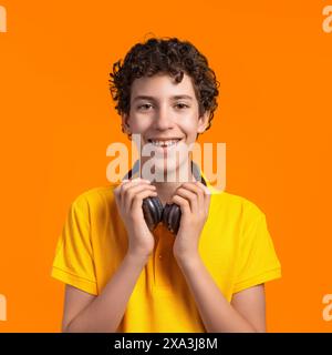 Lächelnder Junge mit lockigem Haar, der Kopfhörer und ein gelbes Hemd trägt, leuchtend orange Hintergrund, selbstbewusstes und ausdrucksstarkes Porträt, Jugendliche Energie und J Stockfoto