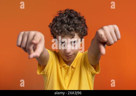 Selbstbewusster Junge mit lockigem Haar, der auf die Kamera zeigt, ein gelbes Hemd, leuchtend orangefarbener Hintergrund, dynamisches und ausdrucksstarkes Porträt, jugendliches Ente Stockfoto