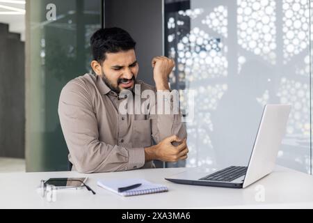 Ein Geschäftsmann leidet bei der Arbeit am Schreibtisch im Büro unter Ellbogenschmerzen, was auf mögliche Belastungen oder Unannehmlichkeiten durch längere Computernutzung hinweist. Stockfoto