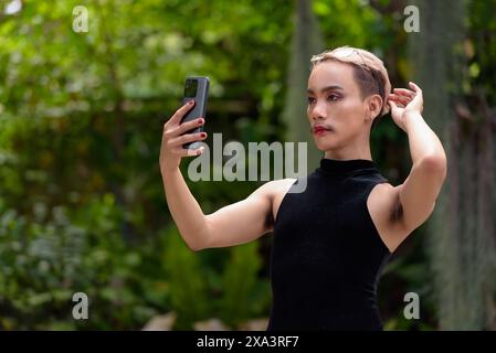 Wunderschöner asiatischer queerer LGBT-Community-Unterstützer Mann mit Schnurrbart, der Lippenstift und Jumpsuit trägt und draußen im Park telefoniert Stockfoto