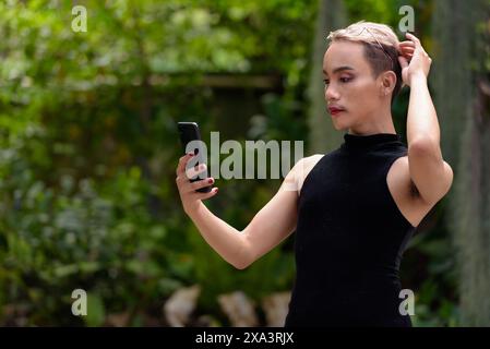 Wunderschöner asiatischer queerer LGBT-Community-Unterstützer Mann mit Schnurrbart, der Lippenstift und Jumpsuit trägt und draußen im Park telefoniert Stockfoto