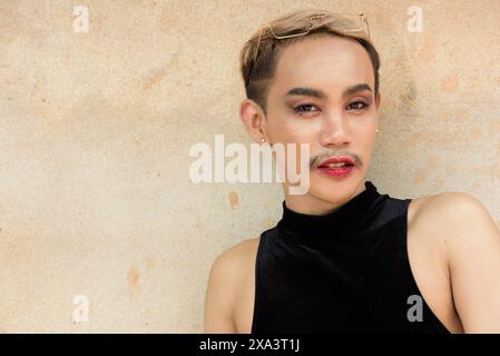 Porträt des schönen asiatischen queeren LGBT Community Unterstützers Mann mit Schnurrbart, der Lippenstift und Jumpsuit vor beigem Wandhintergrund trägt Stockfoto