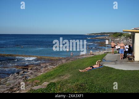 Shelly Beach Ocean Pool, Sonnenanbeter, Pavillon und eine weite Ausdehnung des hellblauen Himmels, blauer Ozean Pool und offenes tiefblaues Meer dahinter, Cronulla Sydney Stockfoto