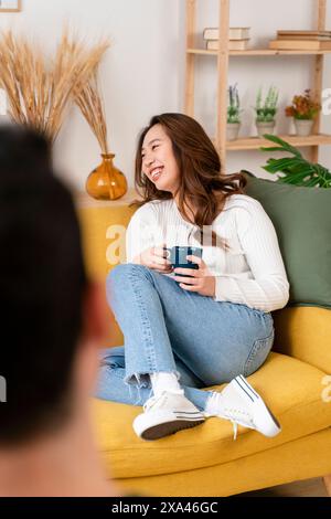 Frau, die auf einem gelben Sofa sitzt und eine Tasse hält und lächelt. Stockfoto