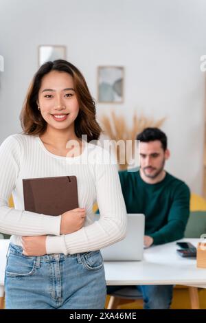 Frau, die mit einem Buch vor einem Mann steht, der an einem Laptop arbeitet. Stockfoto