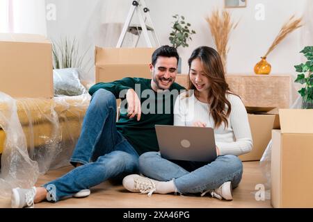Ein Paar lächelt, während er einen Laptop in Umzugskartons benutzt. Stockfoto