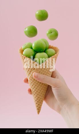 Frauenhand, die Eiskegel mit frischen grünen Pflaumen auf hellem pastellrosa Hintergrund hält. Kreative Komposition für den Sommer. Obst- und Süßigkeitenkonzept. Stockfoto