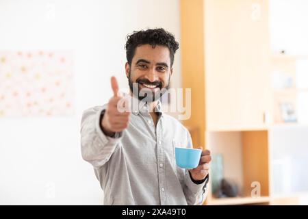Ein lächelnder Mann mit Bart hält einen blauen Becher in einer Hand und zeigt den Daumen hoch Stockfoto